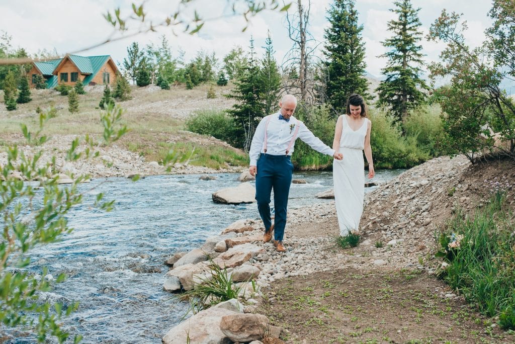 Wedding couple walks along river bank with cabin in the background in Breckenridge, Colorado