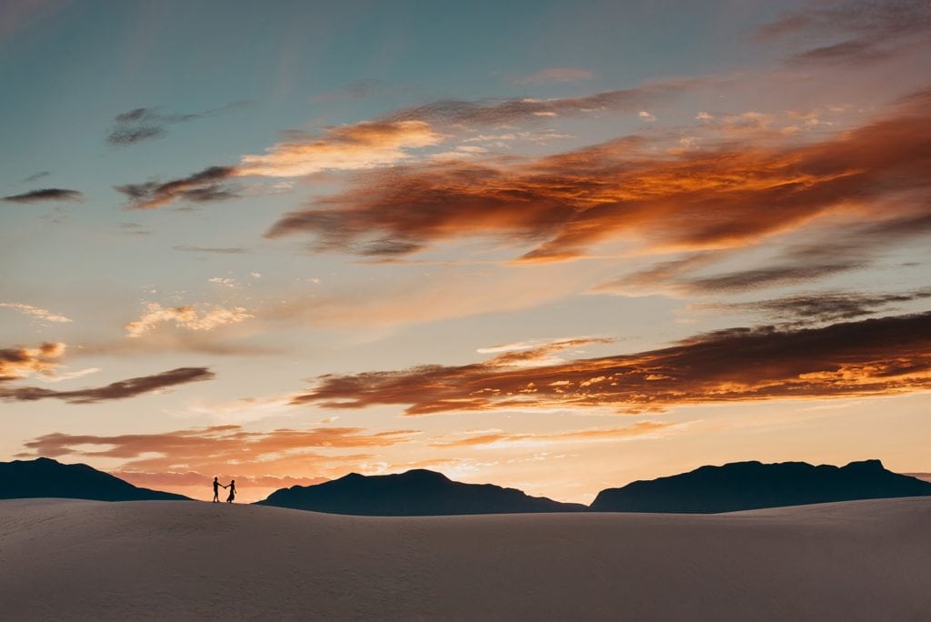 Couple walks across sand dune as silhouettes against sunset with mountains