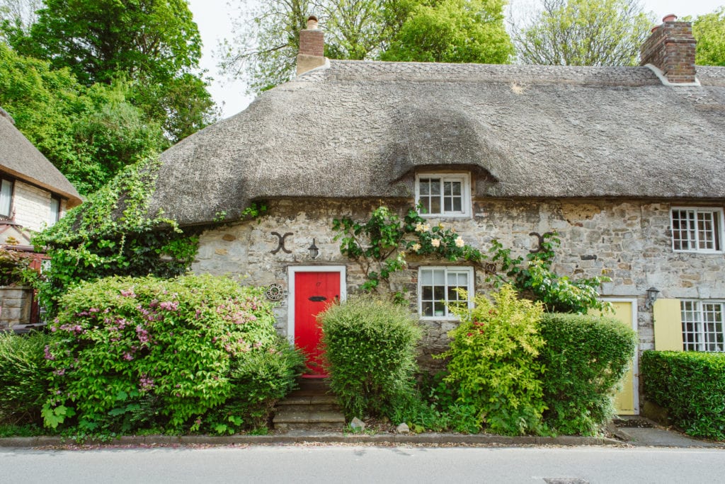 A quintessential English cottage with a thatched roof, vines on the stone walls, and a red door.