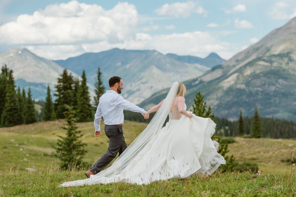 Bride and groom walk hand in hand through grassy hills with big mountains in the background and trees. Bride wears a long cathedral veil and dramatic ballgown wedding dress while groom wears simple dress shirt and dark slacks.