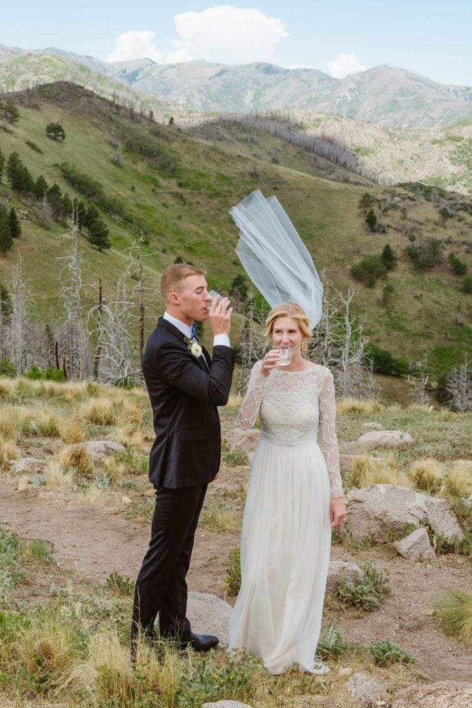 Bride and groom drink champagne on mountain while bride's veil is caught by the wind and stands straight up