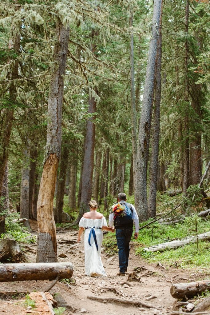 Bride and groom hike forest trail while holding hands. Groom is wearing a backpack with a bouquet sticking out, and bride is wearing an off the shoulder lace dress with blue ribbon belt.