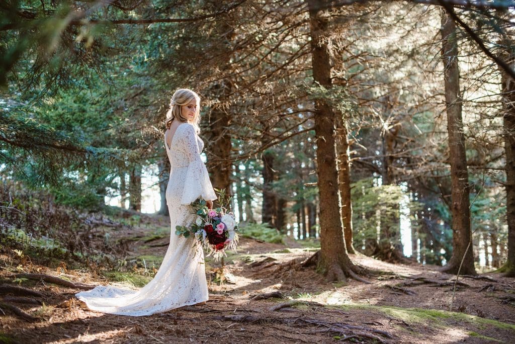 Bride in lace bell sleeve wedding dress stands in sunny forest with her bouquet