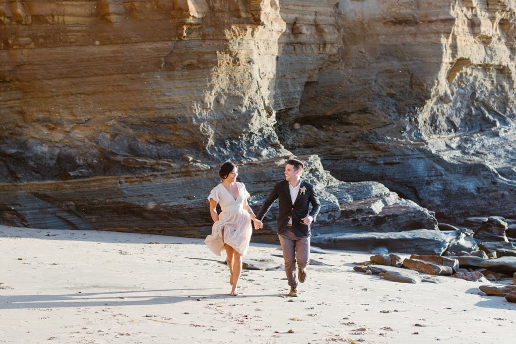 Eloping couple runs down rocky beach while bride holds her dress