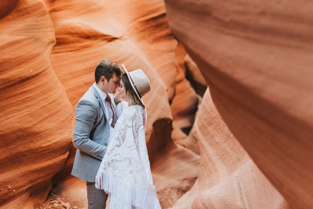 Bride and groom share a sweet moment with their eyes closed and foreheads touching while they're in a red rock slot canyon. Bride wears a Reclamation lace dress and a wide brim hat while groom has a light grew suit jacket and rust colored tie.