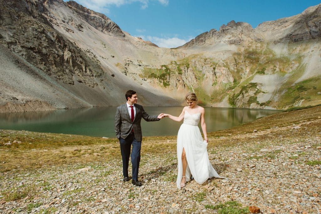 Bride and groom walk along bank of a high alpine lake surrounded by mountains. Bride holds her dress skirt and shows her leg through dress slit