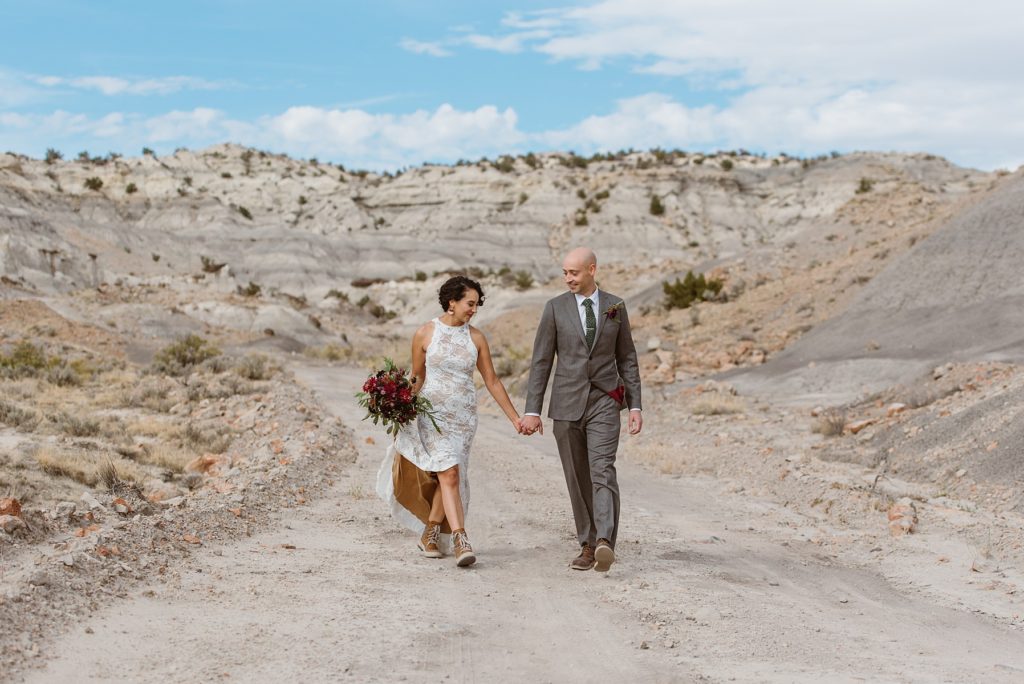 Eloping couple walks down pathway through desert canyon while holding hands. Bride carries her dress train and bouquet.