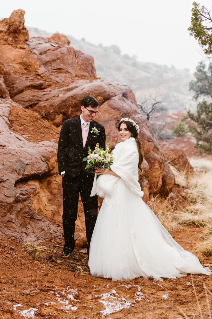 Bride and groom smile while snow falls on them amongst the red rocks on their elopement day. Bride wears a white flower crown and white faux fur stole wrap over her tulle wedding dress.
