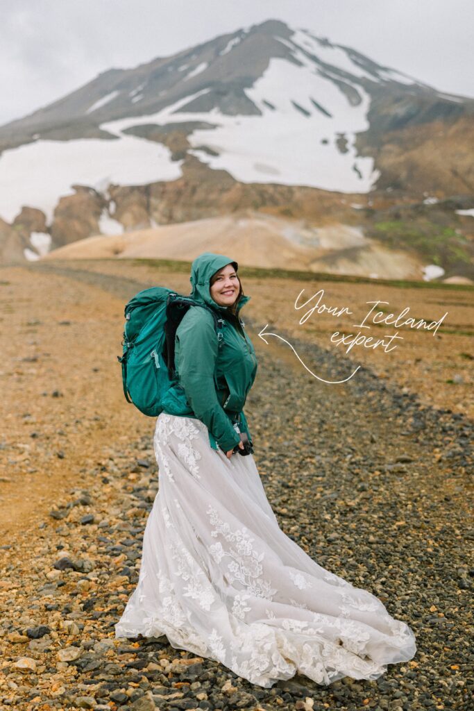 Julie stands on a hiking trail with a snowy mountain in Iceland behind her while wearing a backpack and rain jacket.